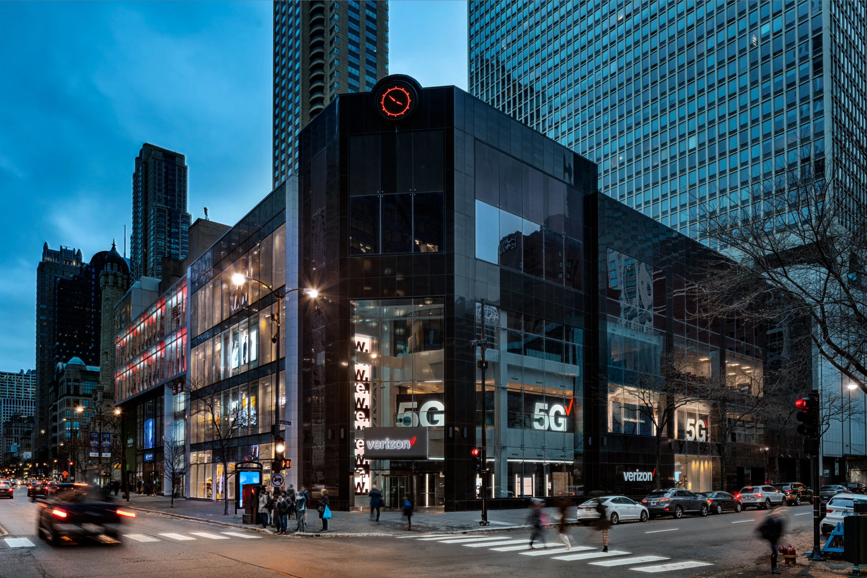 A photograph showcasing Verizon's urban retail experience in Chicago, emphasizing the brand's 5G integration through modern glass architecture and nighttime cityscape.